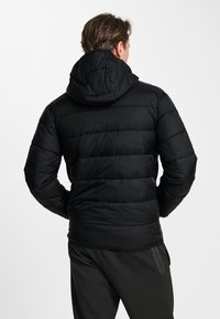 Man die een zwarte gewatteerde jas met capuchon en zwarte broek draagt, gezien van achteren tegen een effen witte achtergrond.