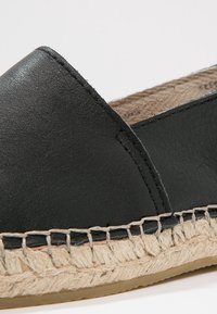 Chaussure espadrille en cuir noir avec semelle en jute tressé beige et coutures visibles, montrant les détails latéraux et de l'orteil.