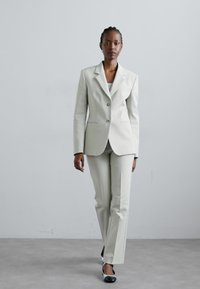 Hellgrauer Anzug mit maßgeschneidertem Blazer, der mit einem Reverskragen und zwei Knöpfen versehen ist, kombiniert mit schmalen Hosen. Schwarz-weiße Schuhe.