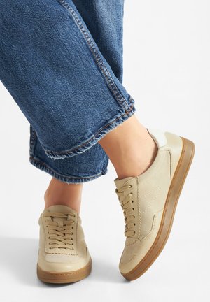 Beżowe trampki wykonane ze skóry i materiału syntetycznego, z płaskimi sznurówkami, gumową podeszwą i białymi akcentami na pięcie.