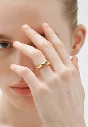 Anillo de oro con una superficie lisa y una pequeña gema incrustada. Mano parcialmente visible, con piel clara y dedos delgados.