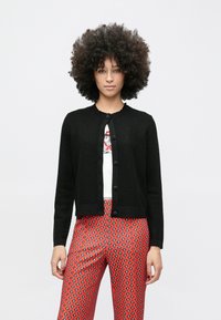 Lokkis juustega naine kannab musta nööpidega kardigani valge graafilise mustriga pluusi peal ning punaste mustriliste pükstega, seistes tavalise valge tausta ees.