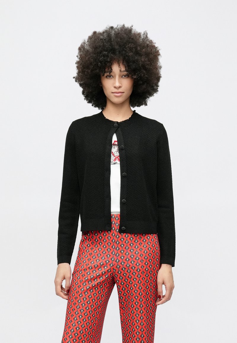 Lokkis juustega naine kannab musta nööpidega kardigani valge graafilise mustriga pluusi peal ning punaste mustriliste pükstega, seistes tavalise valge tausta ees.