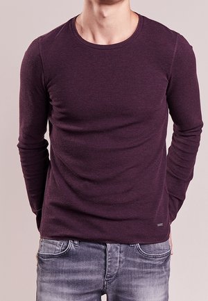 Man met een aansluitend donkerpaars shirt met lange mouwen en vervaagde blauwe spijkerbroek, staand met handen achter de rug.