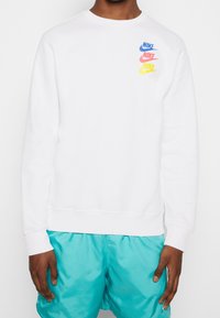 Vit Nike-tröja med rund hals, långa ärmar och en färgglad logga i blått, rosa, gult och orange över bröstet.