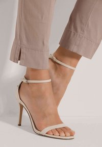 Beige hoge hakken sandalen met dunne bandjes en enkelgesp. Gladde textuur en open teen ontwerp, gecombineerd met lichtgekleurde, cropped broeken.