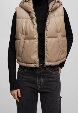Veste sans manches beige matelassée avec capuche portée sur un haut noir à manches longues et un jean noir, modèle debout avec une main dans la poche.