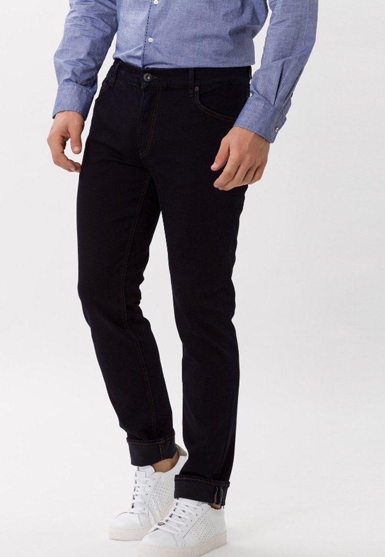 Svarta jeans i denim med orange sömmar, slim fit och uppvikta byxben. I kombination med vita sneakers. Modellen bär en blå skjorta med knappar.