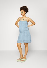 Vestido de ganga azul claro com alças finas e barra com babados. Usado com sandálias de plataforma verdes e óculos retangulares verdes.
