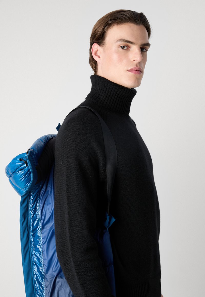 Un pull à col roulé noir tricoté associé à un sac à dos bleu rembourré avec une finition brillante et des bretelles noires étroites.