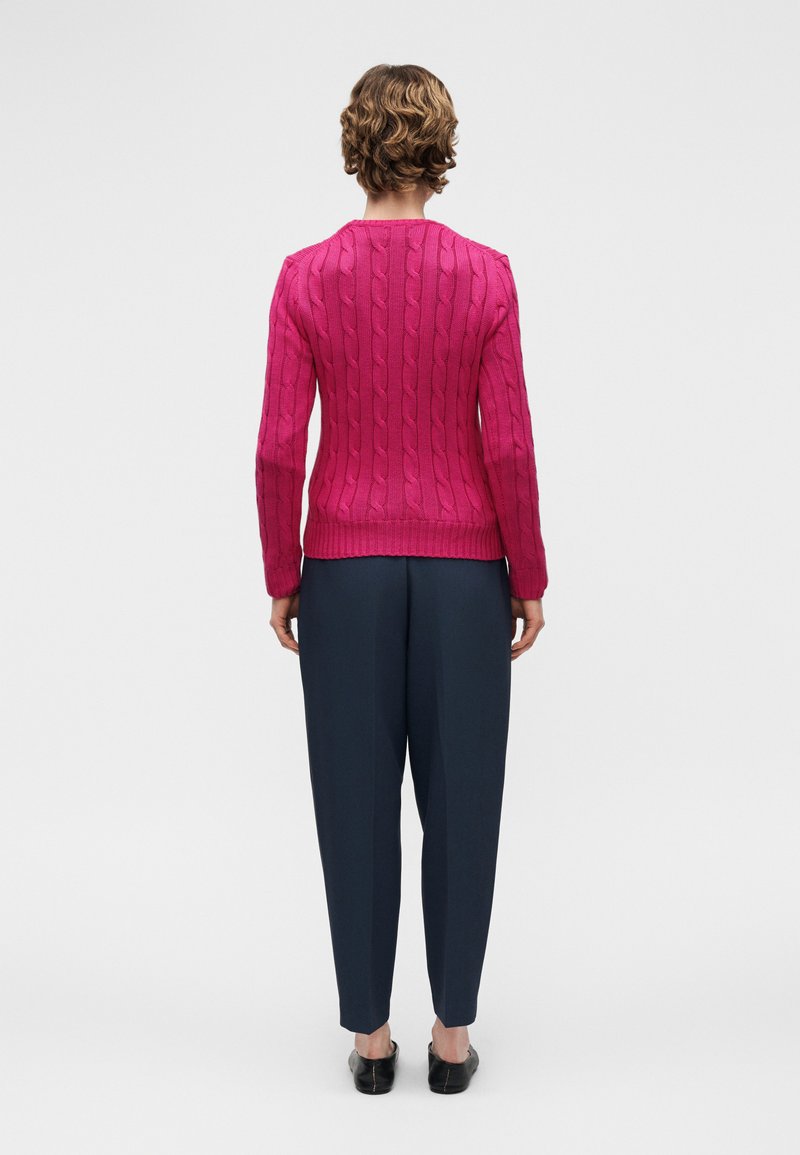 Personne aux cheveux courts et bouclés portant un pull rose ajusté en maille torsadée, un pantalon bleu marine à jambes fuselées et des chaussures noires sans lacets, vue de dos.