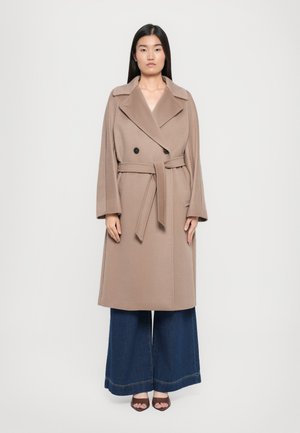 Cappotto beige doppiopetto con cintura, ampi revers e tasche a toppa. Il modello lo indossa sopra a un paio di jeans larghi blu navy e tacchi marroni.