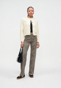 Krémový sveter na zapínanie s okrúhlym výstrihom a zlatými gombíkmi, v páre s hnedo-béžovými nohavicami so vzorom cikcak. Čierna taška a topánky.