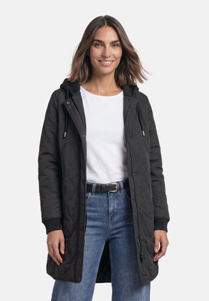 Frau mit schulterlangem braunem Haar, die eine lange schwarze gesteppte Jacke, ein weißes T-Shirt, blaue Jeans und einen schwarzen Gürtel trägt und lächelt.