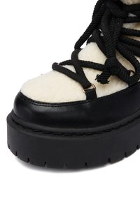 Czarne buty na platformie z miękkim, kremowym wykończeniem z owczej wełny, wyposażone w grube czarne sznurówki i złote akcenty. Teksturowana podeszwa zapewniająca przyczepność.