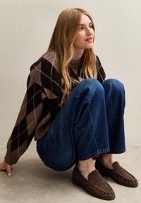 Maglione argyle marrone con collo alto, jeans blu a gamba larga e mocassini marroni. Il maglione presenta un motivo di maglia testurizzata.