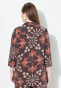 Bunt gemusterte Jacke mit dunklem Hintergrund, auf dem Palm- und geometrische Designs in Orange und Beige zu sehen sind. Dreiviertelärmel.