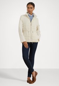 Weißer Puffer-Jacke mit hohem Kragen, gestepptem Design, Frontreißverschluss und Kordelzug in der Taille. Kombiniert mit dunklen Jeans und braunen Slippers.