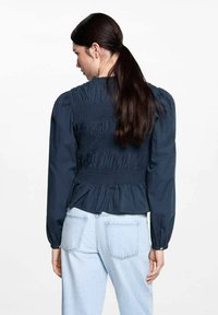 Marinblus med smockad bak, puffärmar och peplumslängd. Tyg av bomull med texturerad finish, bärs med ljusa denimjeans.