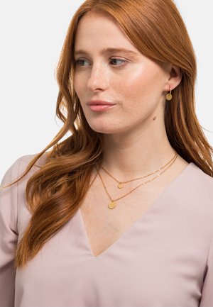 Schmuck aus goldfarbigem Metall mit einem Paar kleiner, runder Ohrringe und geschichteten Halsketten mit runden Anhängern, präsentiert auf einem hellrosa Oberteil.