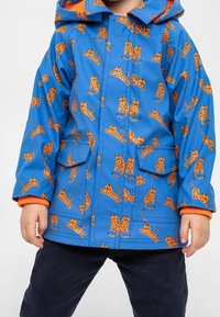 Børns vandtætte jakke i lys blå med orange tiger mønster, der har hætte, to frontlommer og ribbede orange manchetter.