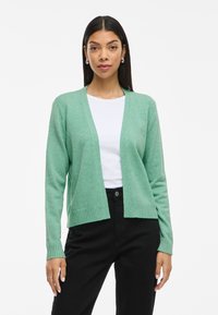 Cardigã verde claro feito de material suave, com decote em V, frente aberta e punhos canelados, combinado com uma blusa branca e calças pretas.