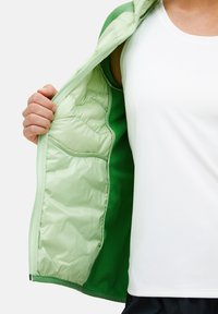 Giacca verde e verde chiaro con zip, dotata di fodera imbottita e tasche laterali, indossata aperta sopra un top bianco. Tessuto morbido e leggero.
