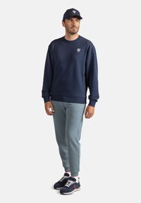 Felpa navy con collo a giro, pantaloni jogger azzurri chiari e sneakers navy con dettagli bianchi. Presenta un logo sul petto e un cappellino. Abbigliamento sportivo casual.