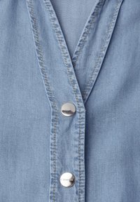 Nærbillede af lyseblåt denimstof med beige syning, med en V-hals søm og to sølvfarvede metaltrykknapper mærket "Street One."