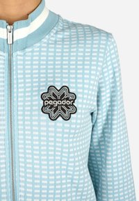 Svetlomodrá bunda s textúrovaným štvorcovým vzorom, na ktorej je čierne vyšívané logo hviezdy s nápisom "pegador" v strede.