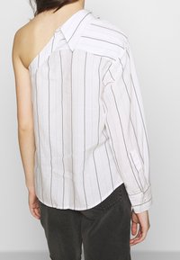 Camisa blanca con rayas verticales marrones, un hombro al descubierto, cuello y manga larga en el lado opuesto, tela ligera y crujiente.