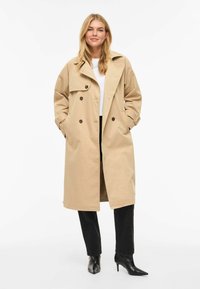 Trench-coat beige avec boutons croisés, large col et poignets à ceinture. Associé à un pantalon noir et des talons noirs pointus.