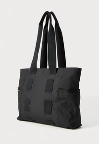 Svart tote bag laget av slitesterkt stoff med to lange håndtak, med en jevn tekstur, flere vertikale paneldetaljer og en sidelomme.