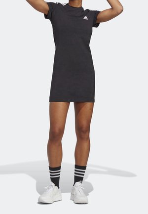 Persoon draagt een zwarte Adidas t-shirt jurk, zwarte sokken met witte strepen en witte sneakers, staat met opgeheven armen.