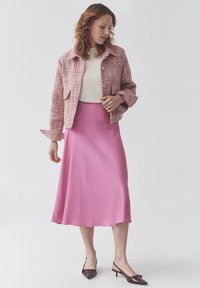 Chaqueta de tweed rosa con dos bolsillos frontales, combinada con una blusa de color claro y una falda larga y fluida rosa. Zapatos de charol con punta afilada y tira al talón.