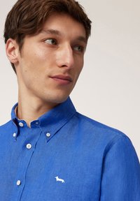 Camisa de lino azul con collar abotonado, con botones blancos y un pequeño logo bordado de un dachshund en el pecho.