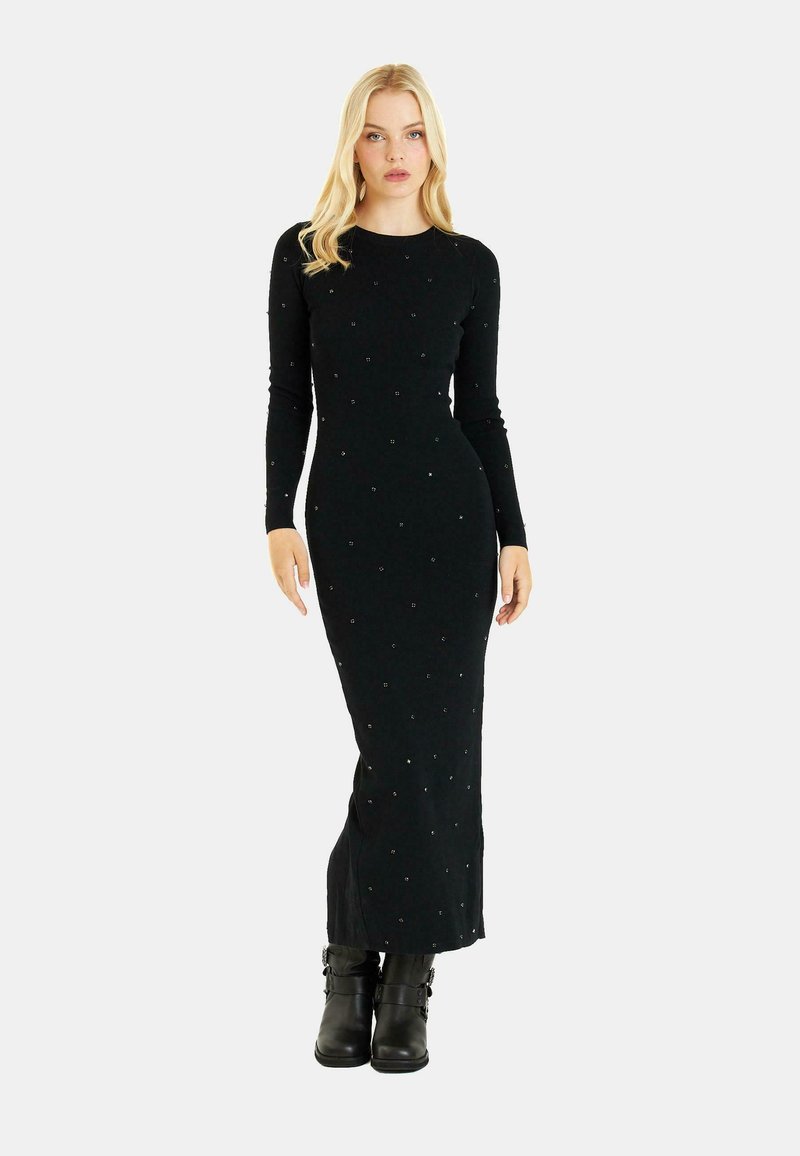 Schwarzes geripptes Maxikleid mit langen Ärmeln, verziert mit verstreuten dekorativen Akzenten und einer taillierten Silhouette, kombiniert mit schwarzen Stiefeletten.