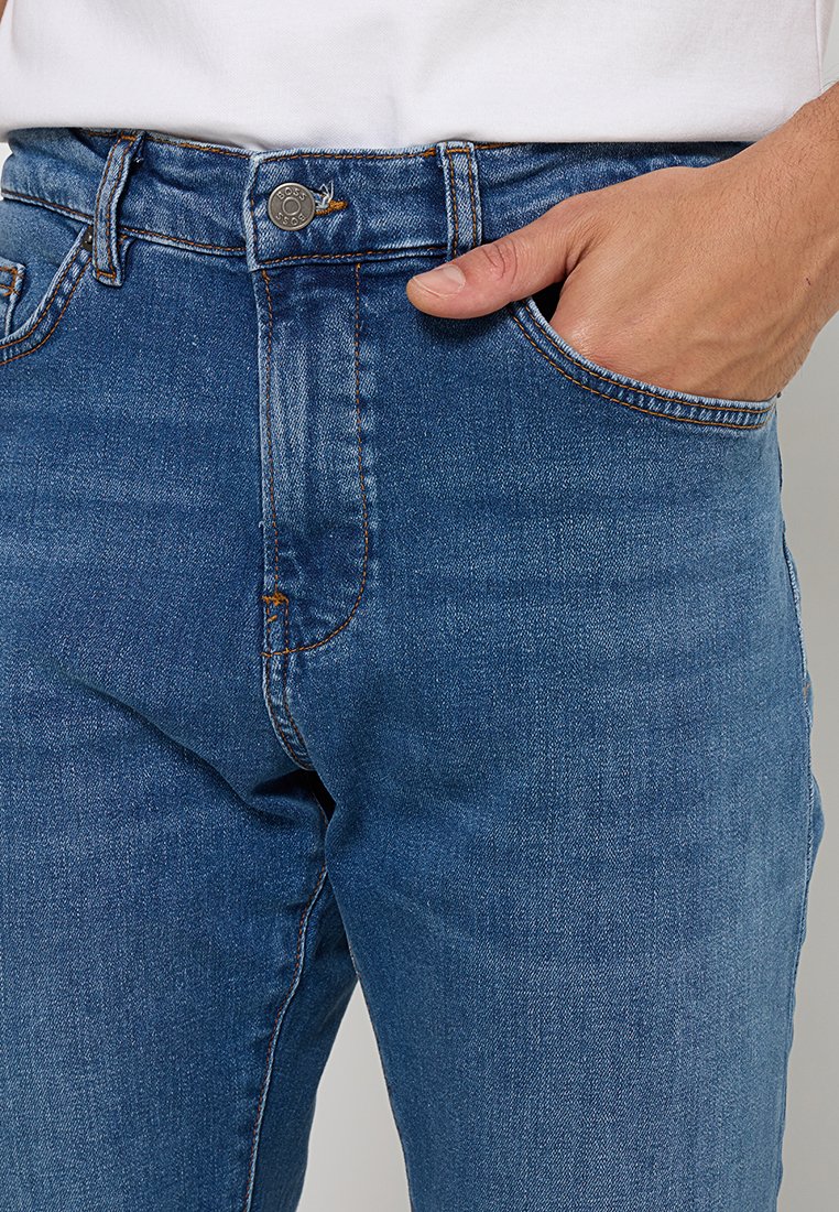 Niebieskie dżinsy o prostym kroju, z widocznymi szwami, przednią kieszenią i metalowym zapięciem na guzik w talii.