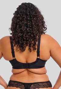Reggiseno in pizzo nero con spalline regolabili, chiusura posteriore e fascia texturizzata, abbinato a mutandine in pizzo nero coordinate, con capelli ricci in vista.