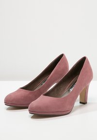 Ein Paar staubige rosa Wildlederschuhe mit geschlossenen, abgerundeten Zehen und Blockabsätzen auf einer weißen Oberfläche.