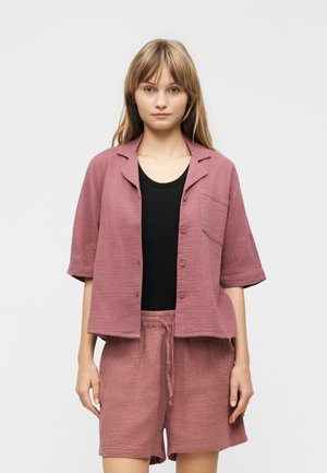 Ung kvinne iført løs mauve knappet jakke og matchende shorts over en svart topp, stående mot en ensfarget lys bakgrunn.