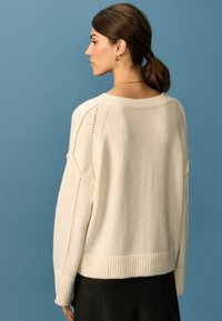 Cremefarvet strikket sweater med ribbestrikket kant, brede ærmer og åbne strikdetaljer. Bagfra set mod en blå baggrund.