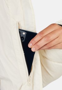 Giacca puffer bianca con tasca con zip, che mostra uno smartphone nero parzialmente inserito. Texture liscia con una leggera lucentezza.