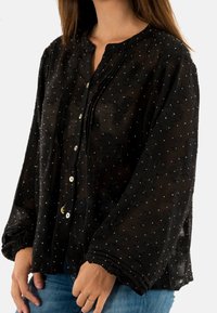 Blusa negra transparente con bordados florales y detalles de lentejuelas, frente con botones y mangas sueltas con puños fruncidos.