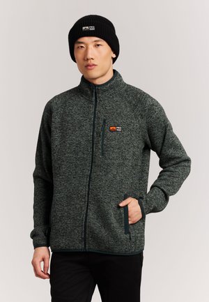 Joven con chaqueta de forro polar gris oscuro con cremallera y gorro negro con logotipos "PRO TEST", posando con una mano en el bolsillo de la chaqueta.