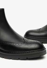 Bottines en cuir noir avec un bout arrondi, présentant des perforations brogue et une semelle texturée, conçues avec une silhouette moderne et élégante.