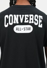 Juoda medvilninė marškinėlių su balta "CONVERSE" užrašu ir ovalo formos "ALL★STAR" logotipu ant nugaros. Standartinio silueto, su trumpomis rankovėmis.