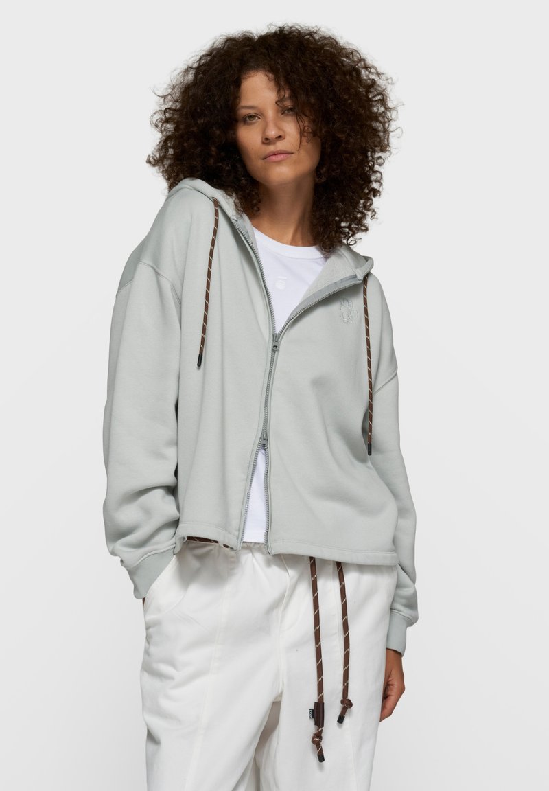 Mujer con cabello rizado que lleva una sudadera con capucha gris clara con cremallera y cordones marrones, camiseta blanca y pantalones blancos, frente a un fondo liso.