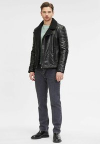 Giubotto in pelle nera con colletto in shearling, dettagli a costine sulle spalle, abbinato a una camicia verde chiaro e jeans grigi scuri. Scarpe nere.