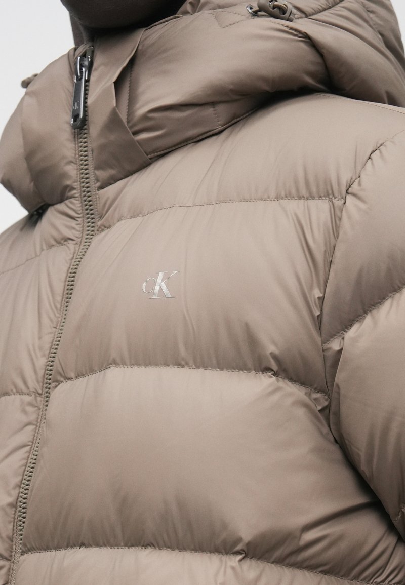 Braune gesteppte Jacke mit Reißverschluss und Kapuze, mit einem kleinen silbernen "cK"-Logo auf der Brust.